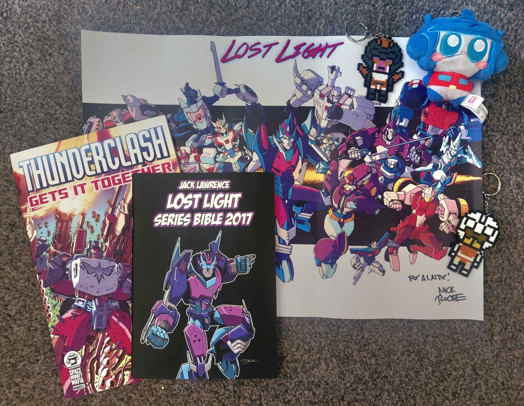 A Lost Light art print by Nick Roche, a comic titled 'Thunderclash Gets it Together', Jack Lawrence's 2017 Lost Light Series Bible, keychains of Aileron, Chromedome, and Optimus Prime, all in a loose pile on grey carpet.